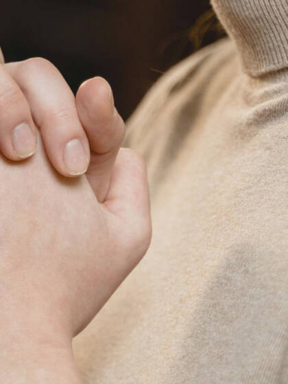Kaksi kättä on ristissä, toinen käsi on päällä toista. Taustalla näkyy epämääräinen sisustus.